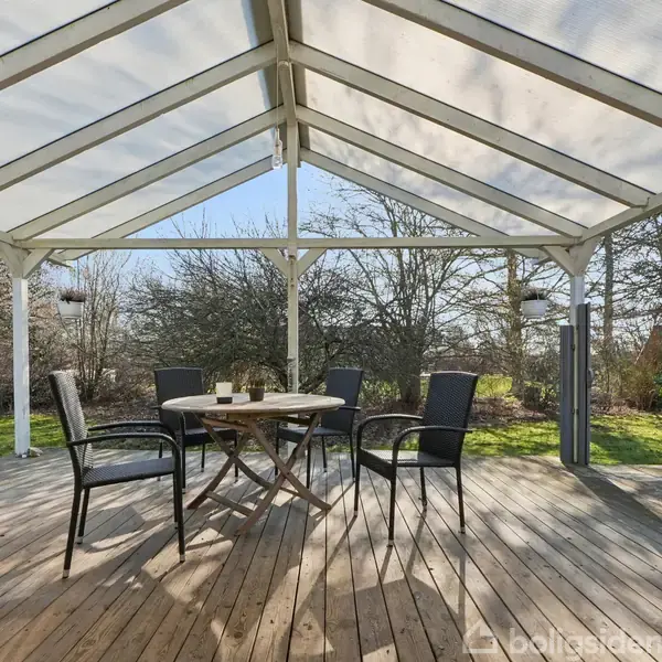Terrasse med trægulv og gennemsigtigt tag, fem stole omkring et rundt bord. Omgivet af græsplæne og træer, skaber en rolig atmosfære.