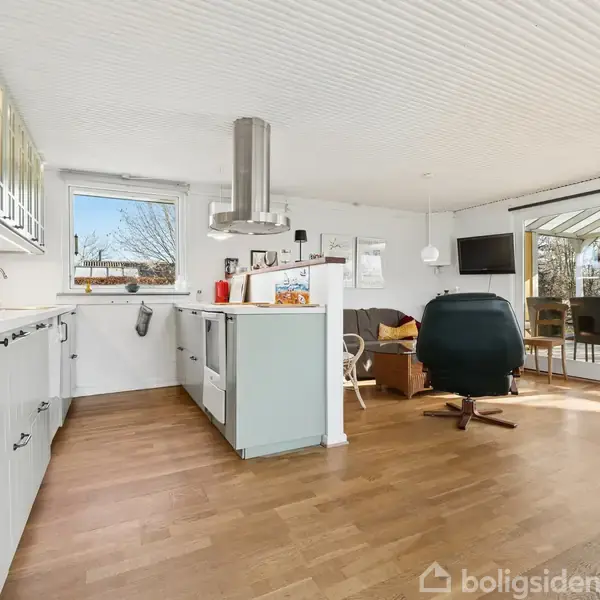 Et moderne køkken med grønne skabe står i et lyst, åbent rum. En kakkelovn, en sort lænestol og en spiseplads ses i baggrunden. Store vinduer giver udsigt til en terrasse.
