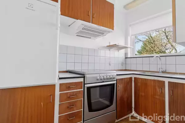 Komfur med ovn står i et hjørnekøkken, flankeret af skabe i træfinish og en hvid Electrolux køleskab. Over komfuret er en emhætte. Et vindue over vasken viser træer udenfor.