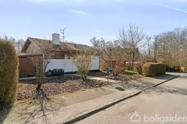 Hus med hvid facade og skråt tag står på en stille vej. Omgivet af lave buske og små træer uden blade i en forstadslignende atmosfære.