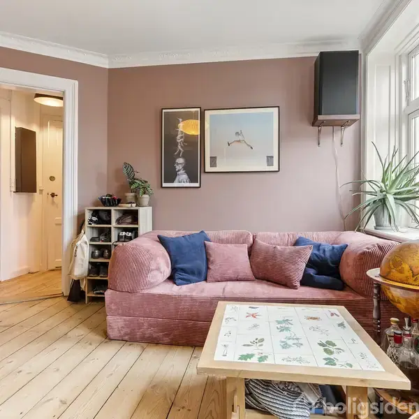 En lyserød sofa med puder står ved vinduet i en stue med parketgulve. Et rundt globus-barskab og en trækommode ses ved siden af planter og kunstværker på væggene.