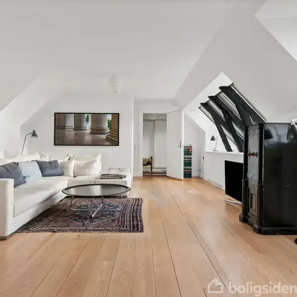 En lys stue med en hvid sofa ved siden af et sofabord på et tæppe. Store skråvinduer over stuepladsen. Trægulve og en sort brændeovn ved siden af.