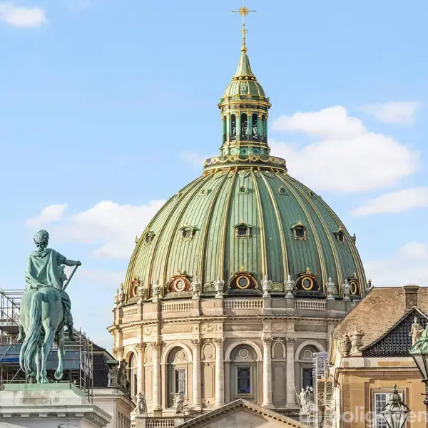 Kuplen står majestætisk over bygningen, dekoreret med grønne paneler og ornamenter, med en skulptur til venstre. Bygningen er i klassisk stil, himlen er klar med få skyer.