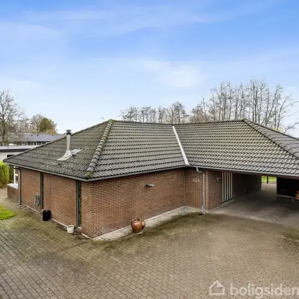 En brun murstensvilla med gråt tegltag står på en flisebelagt indkørsel. En carport er integreret i huset. Omkring er der træer og en blå himmel.