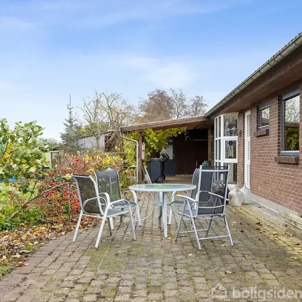 Terrassemøbler står på en stenbelagt havegang ved siden af et murstenshus. Fire stole omringer et lille bord. Haven er omgivet af vegetation og træer i baggrunden.
