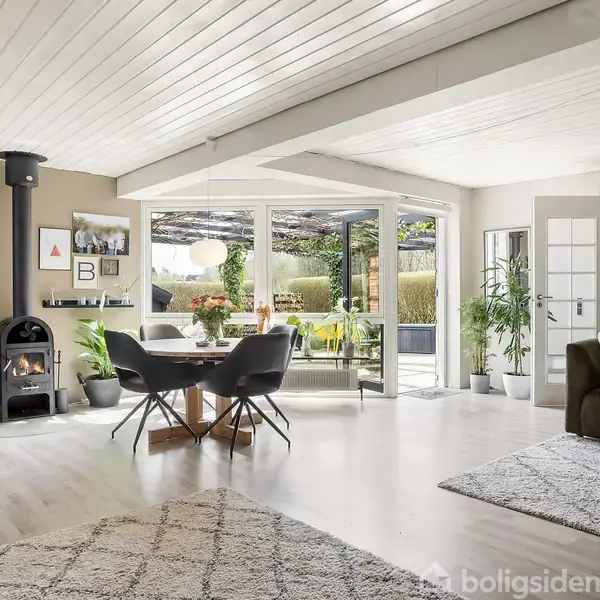 Et lyst, moderne stueinteriør med en pejs, omkring hvilken står en lænestol. En spiseplads med runde stole er placeret nær store vinduer, der åbner mod en terrasse og have.