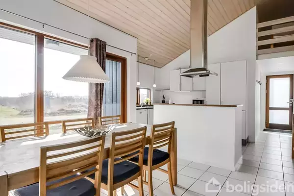 En lys spisestue med et træbord og seks stole, oversvømmet af sollys fra store vinduer. Køkkenet i baggrunden har hvide skabe og en emhætte.