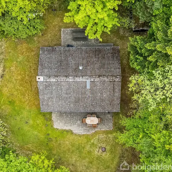 En firkantet bygning med et skråt tag set ovenfra. Bygningen har en terrasse med et rundt bord. Omgivet af tæt grøn vegetation og en græsplæne.