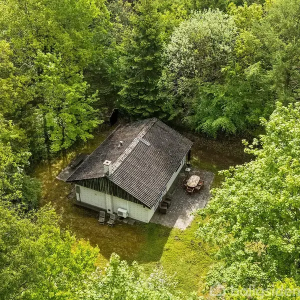Et hus ligger stille blandt tætbevoksede grønne træer. På en terrasse ved siden af huset står havemøbler. Omgivelserne er frodige og skovklædte.