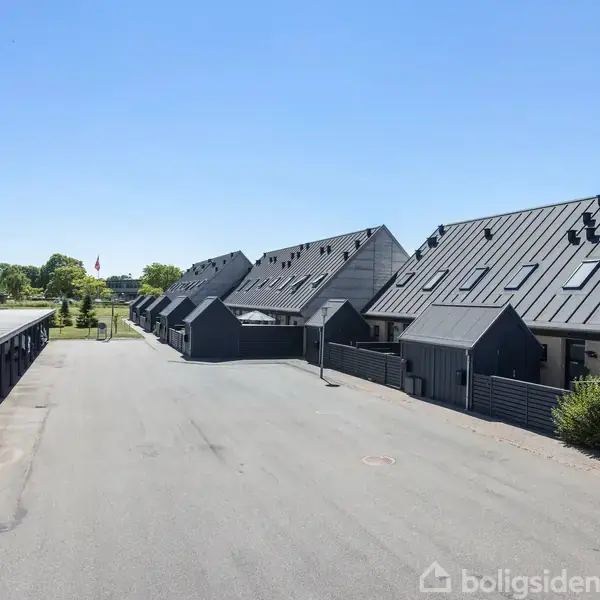 Rækkehuse med skrå tage står på række langs en asfalteret vej, omkranset af carporte til venstre. Solrigt og grønt område i baggrunden.