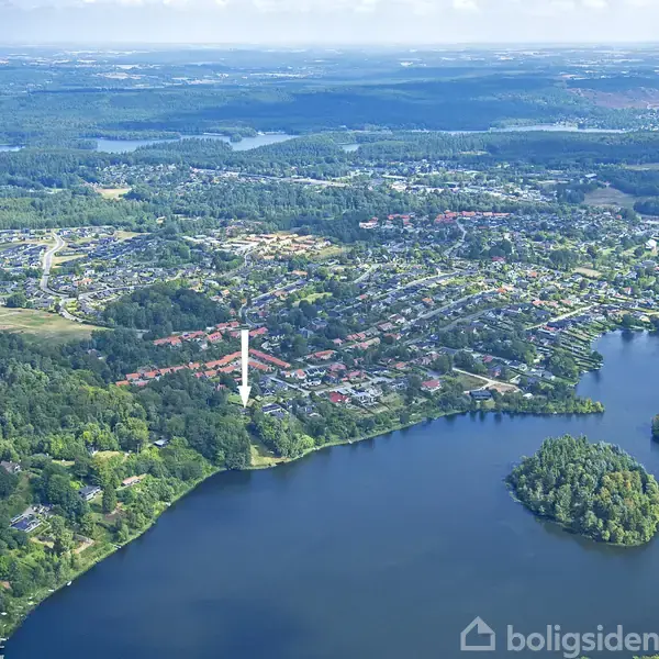 Flyfoto viser en sø omgivet af skove og bygninger. En pil peger mod et bestemt område, der er tæt pakket med huse. Området ligger i et grønt, kuperet landskab.