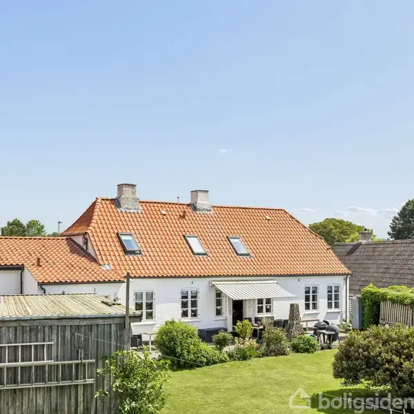 Hvidt hus med rødt tegltag og flere vinduer, står i midten af en solrig have omringet af hække og træer. Tre skorstene hæver sig fra taget, og solen skinner klart.