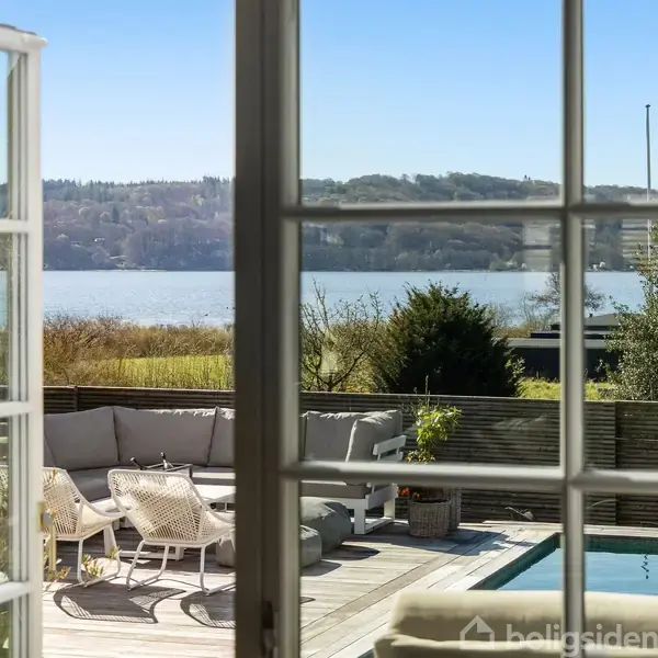 Glasdør åbner ud til en terrasse med sofa og stole, ved siden af en pool. I baggrunden ses en sø og bakkede landskaber under en klar blå himmel.