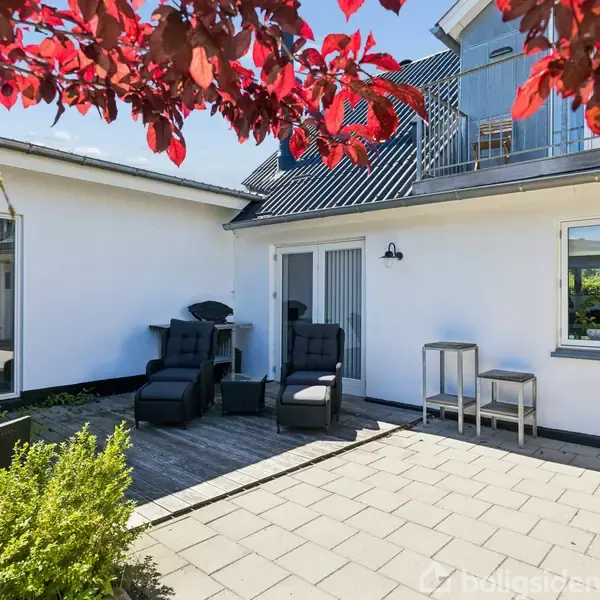Hvide havemøbler står på en solrig terrasse foran et moderne hvidt hus. Omkring er der grønne buske og et træ med røde blade, der tilføjer farve til omgivelserne.