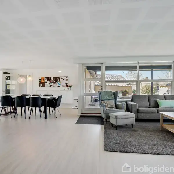 Stue med grå hjørnesofa, sofabord og lys trælaminatgulv. Et spisebord med sorte stole står nær åben hvidt køkken. Store vinduer giver udsigt til træer udenfor.