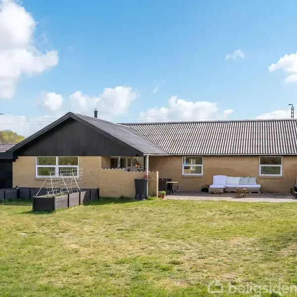 Hus med fladt tag i ét plan, beige murstensfacade, vinduer og terrasse, omgivet af stor græsplæne. Blå himmel med spredte skyer.
