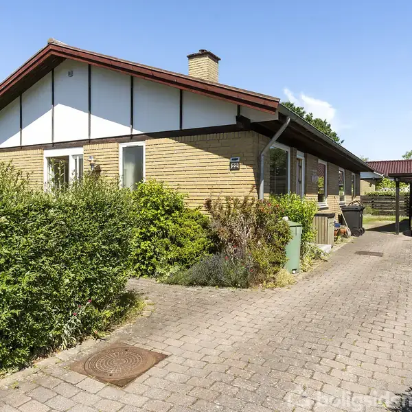Et enfamiliehus i et boligområde har en hældende tagkonstruktion. En bil holder i en carport, og en brolagt indkørsel omgives af grønne buske.