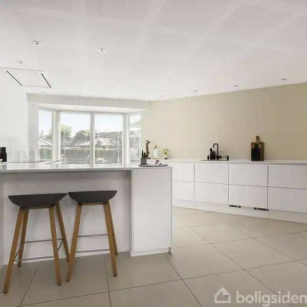 Moderne køkken med hvide skabe og bordplader, to barstole ved en ø. Store vinduer bringer naturligt lys ind, mens planter og køkkenudstyr pynter bordpladen. Beige vægge skaber en varm atmosfære.