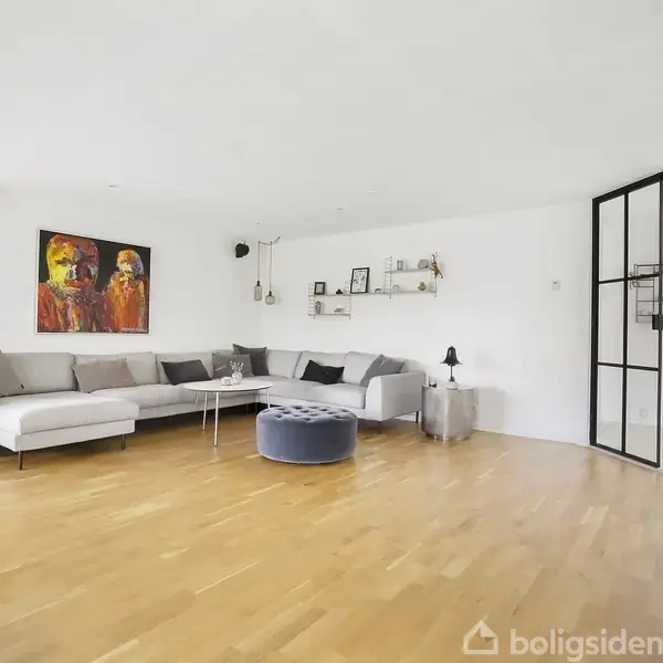 Stor stue med hvid L-formet sofa, lille rundt bord og lampe. Moderne kunst hænger på væggen ved siden af reoler. Rummet har trægulv og glasvægsafdeling.