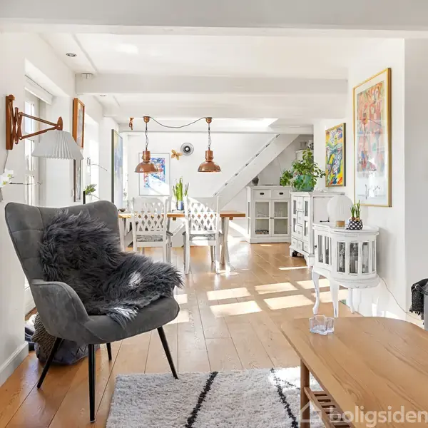 En lys stue med moderne indretning; en grå lænestol med pude og tæppe står foran et tv. Et hvidt spisebord med stole ses i det solfyldte baggrundsrum.