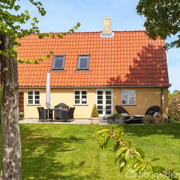 Et gult hus med rødt tag står i en have. Der er en udendørs spiseplads med parasol foran huset og grønne buske omkranser plænen.