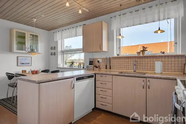 Et lyst køkken med lyse træskabe og grå bordplader. En opvaskemaskine er indbygget. Køkkenvinduerne giver udsigt til en rød tagryg. Der hænger lamper over bordet og vasken.