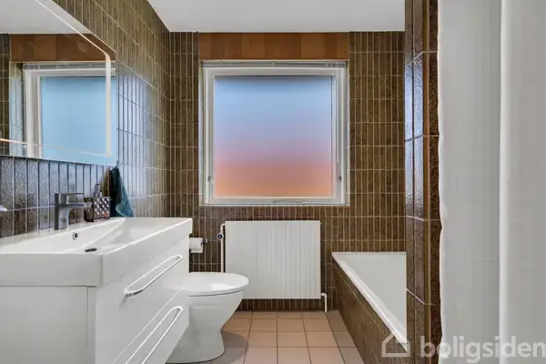 Et hvidt toilet og håndvask står mod en brunt flisebelagt væg. Et vindue med frostet glas viser en gradient af blå til rosa. En radiator og badekar er ved siden af.