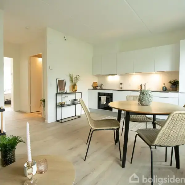 Spisebord med fire stole står i en moderne, lys stue-køkken kombination. Køkkenet oplyses af indbyggede lamper, og stuen har planter og dekorationer på borde og hylder.