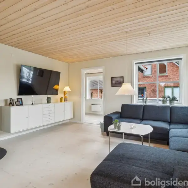 En stor, mørkegrå sofa står i en lys stue med træloft. Tv'et hænger over en hvid skænk ved siden af et brændekomfur. Store vinduer giver udsigt til røde murstenhuse.