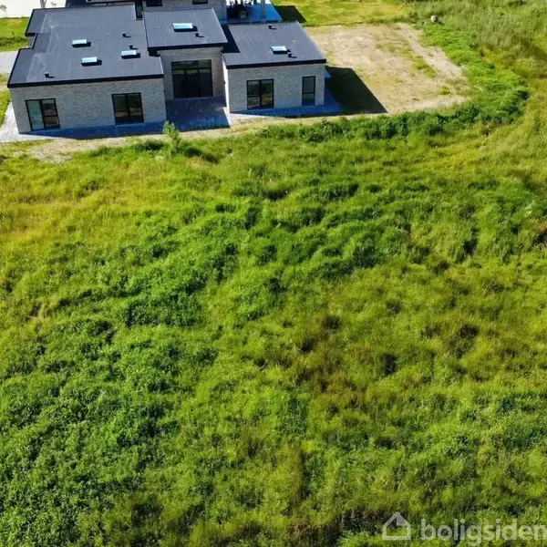 Et moderne hus med fladt tag står stille, omkranset af en stor, grøn græsplæne. En vej løber tæt forbi i venstre side.