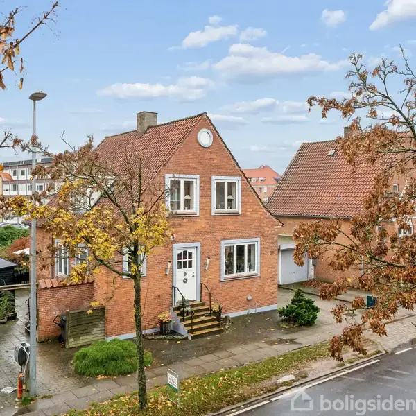 Rødt murstenshus med hvide vinduer står ved en fortovskant. Trappetrin fører til indgang, omkranset af efterårstræer. Gade og naboer ses i baggrunden under en blå himmel med skyer.