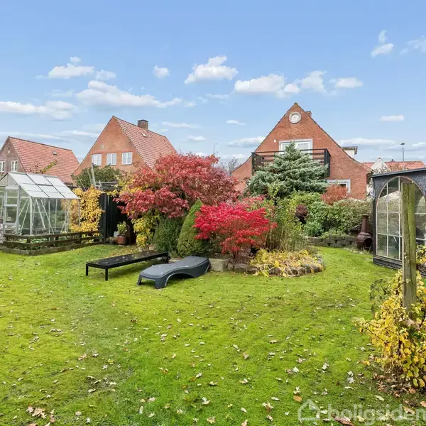 Et hus med rødt tegltag står bag en frodig have med grønne plæner, farverige buske, et drivhus og en pavillon. Himlen er klar med spredte skyer.