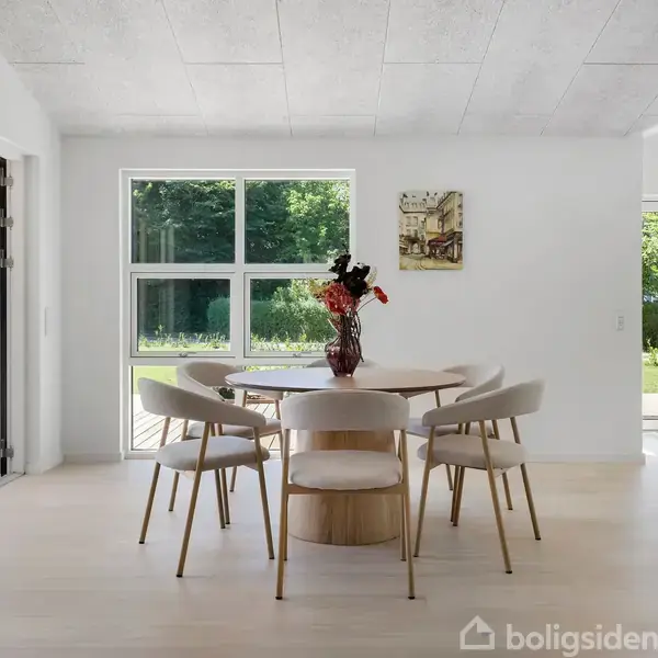 Spisestue med gråt hjørnesofa står ved siden af et geometrisk tæppe. Et spisebord med stole er placeret nær vinduer med udsigt til haven. Kunst pryder væggen.