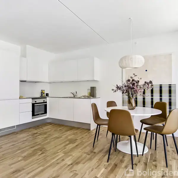 Et hvidt, moderne køkken med integrerede apparater og en rund spisebord med brunlæder stole. En pendellampe hænger over bordet, og vaser med grene står på bordet.