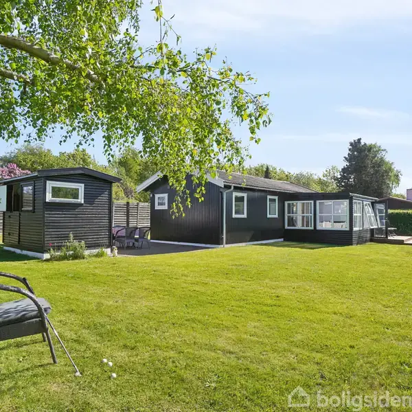 Sort sommerhus med store vinduer står på en velplejet grøn græsplæne. En sort stol med armlæn er placeret i haven, omgivet af træer og buske under en blå himmel.