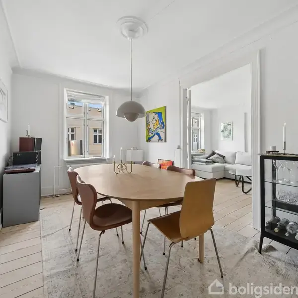 Et rundt spisebord omgivet af stole står i et lyst rum. En moderne lampe hænger over bordet. Væggene har kunst og vinduet viser udsigt til bygninger udenfor.