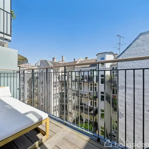 En balkon med en hvid sofa står stille omgivet af flere bygninger. Det er en solrig dag, og himlen er klar blå. Balkonen har sort rækværk.