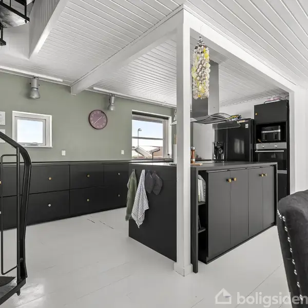 En sort metaltrappe fører opad i et moderne køkken med sorte skabe, hvide vægge og loft. En spisebordstol står til højre, og et ur hænger på væggen.