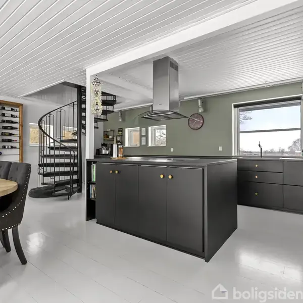 Sort køkkenbord med emhætte over, omgivet af spisebord med polstrede stole. I baggrunden ses en vindeltrappe og et vinreol i en lys, moderne stue med grønne vægge.