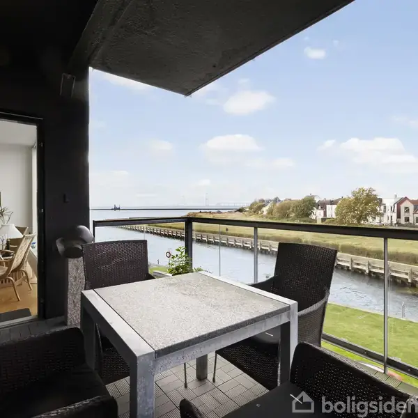 En altan møbleret med et hvidt bord og fire sorte stole. Den vender ud mod en kanal omgivet af græs og huse. En dør åbner ind til en stue.