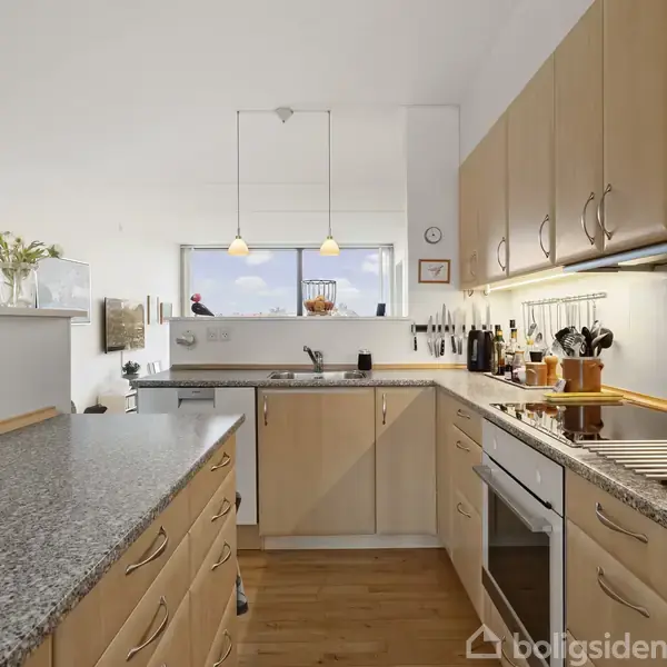 Et lyst køkken med lyse træskabe og granitbordplade. Køkkenet har moderne apparater, hængende lamper og udsigt ud over en lys stue. Reoler med bøger og dekorationer ses i baggrunden.