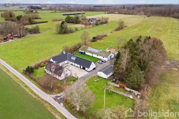 En landbrugsejendom med flere bygninger står samlet midt i omgivende grønne marker og træer. En smal vej fører forbi ejendommen, som er omgivet af åbne landskaber og spredte træer.