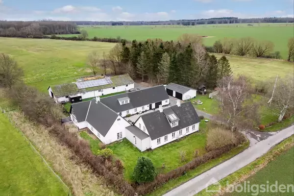 Gårde, som består af flere hvidmalede bygninger med mørkt tag, ligger i et grønt og landligt område omgivet af marker og skove.