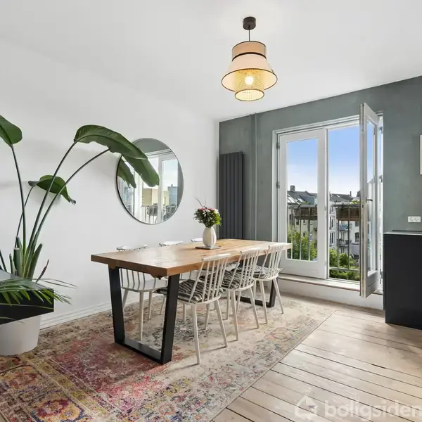 Spisebord med stole står på tæppe, omgivet af grønne planter. En rund vægspejl hænger ved siden af åbne døre, der fører til balkon med udsigt over byens tage.