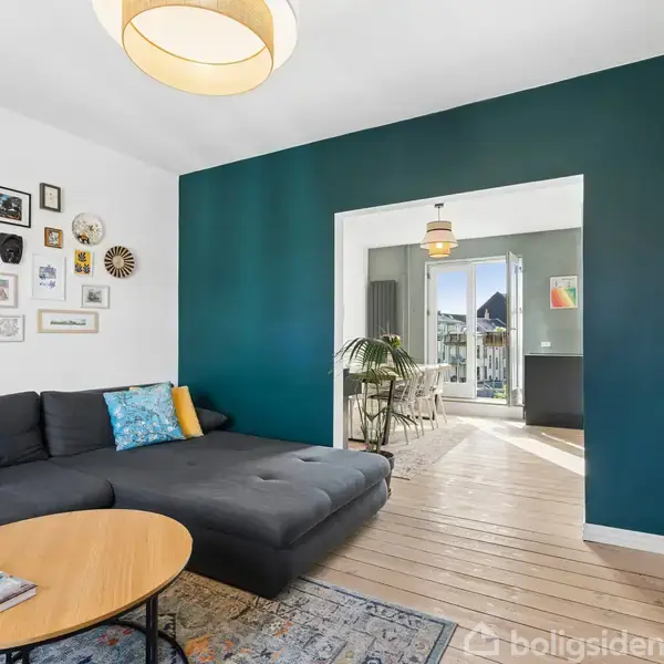 En mørkegrå sofa med farverige puder står i en stue, der har hvide vægge dekoreret med kunstværker. Sofaen står ved et rundt sofabord. Baggrunden viser et åbent rum med spisebord.