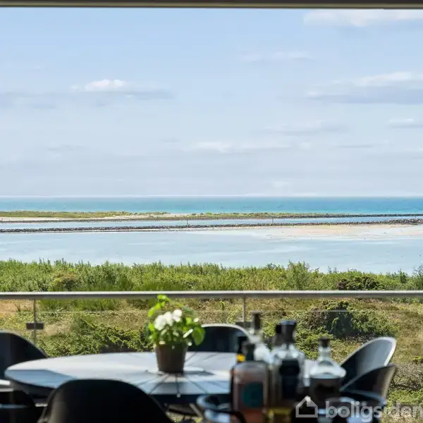 Et rundt bord med stole står på en terrasse med en plante som centrum. Baggrunden viser udsigt over grøn vegetation, vand og horisonten under en blå himmel med skyer.