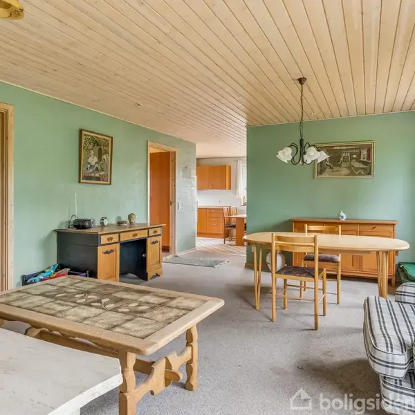 Et rum med træloft og grønne vægge. Der står et spisebord med stole og et sofabord. En dør fører til et køkken i baggrunden.
