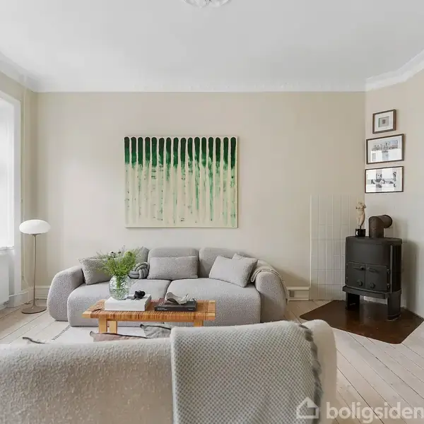 En stue indeholder en grå sofa med tæppe, sofabord og planter. Et maleri hænger på væggen. En brændeovn står ved en TV-skænk. Store vinduer lader lys ind.