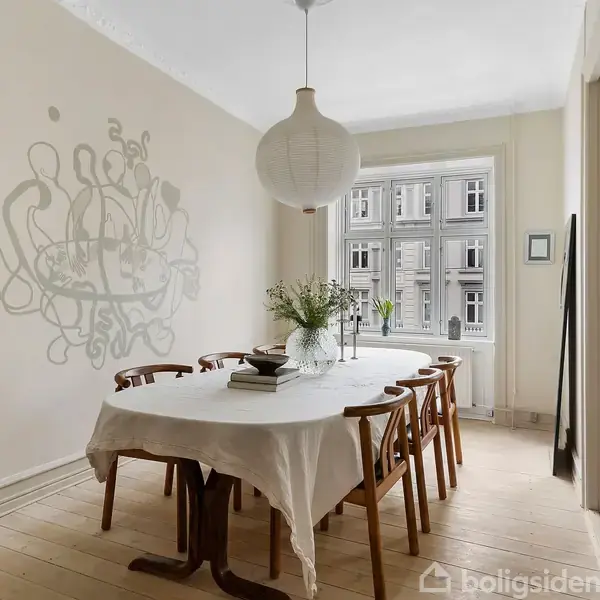 Langbord dækket med hvid dug og omgivet af træstole står i et lyst rum. En stor pendel hænger over bordet, og en vase med blomster pynter midten. Vægmaleri til venstre, vindue til højre.