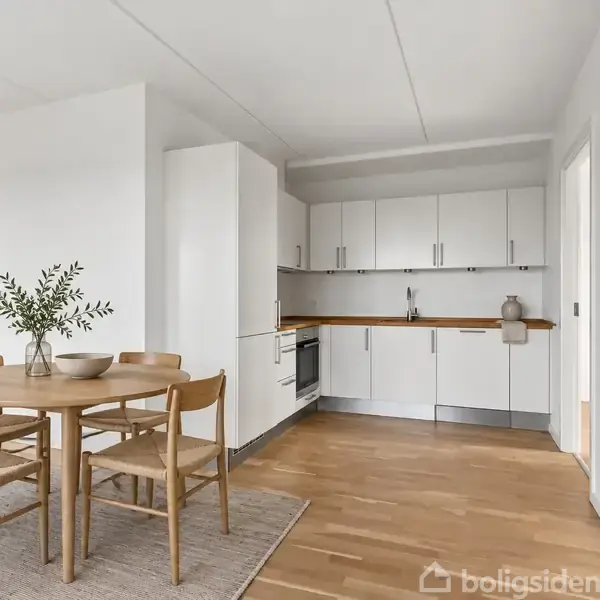 Et spisebord med fire stole står på et tæppe. Køkkenelementer og apparater er i baggrunden i et lyst og moderne rum med trægulv. En plante i en vase pynter på bordet.